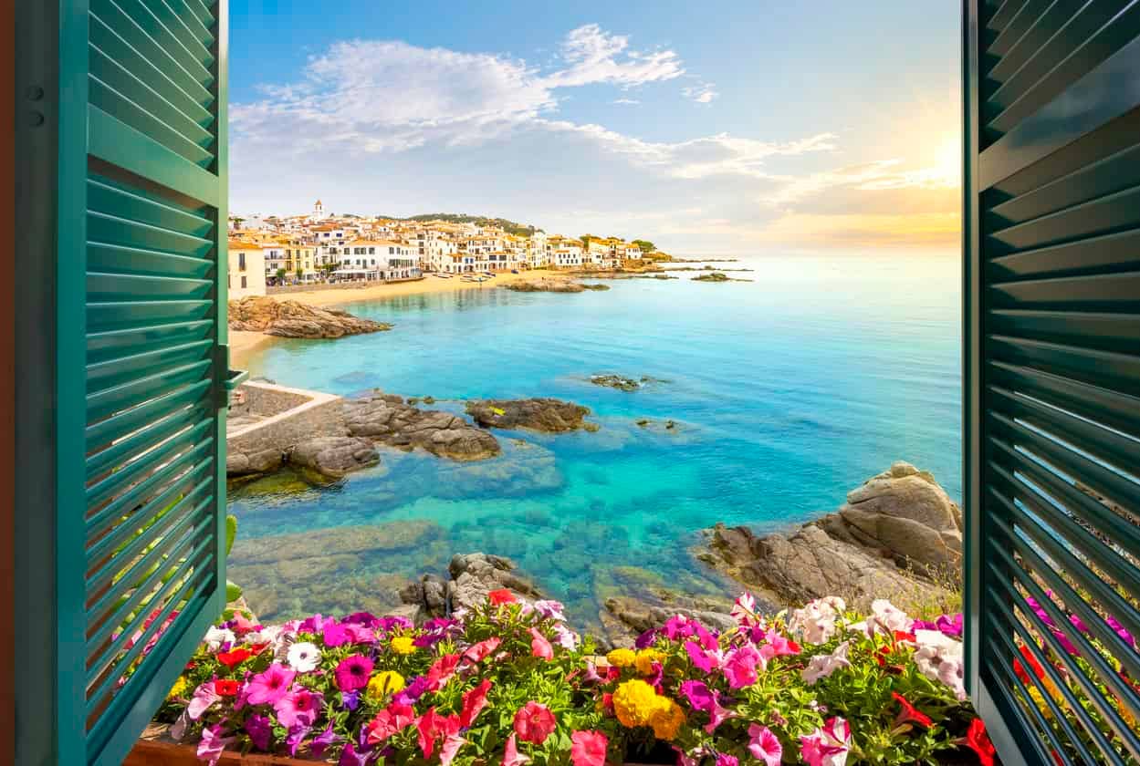 View through an open window with shutters of the sandy beach, rocky coastline and whitewashed town of Calella de Palafrugell, Spain, on the Costa Brava coast as the sun sets. The best summer activities for kids in the Costa Brava.