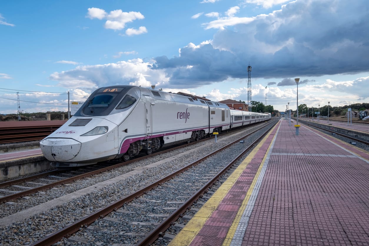 A modern train stopped at the old and still functioning Monfrague or Palazuelo Empalme train station. How to book a train in Spain using Trainline vs Omio, comparing whether Omio train is reliable and if Trainline or Omio is better for Spanish rail travel