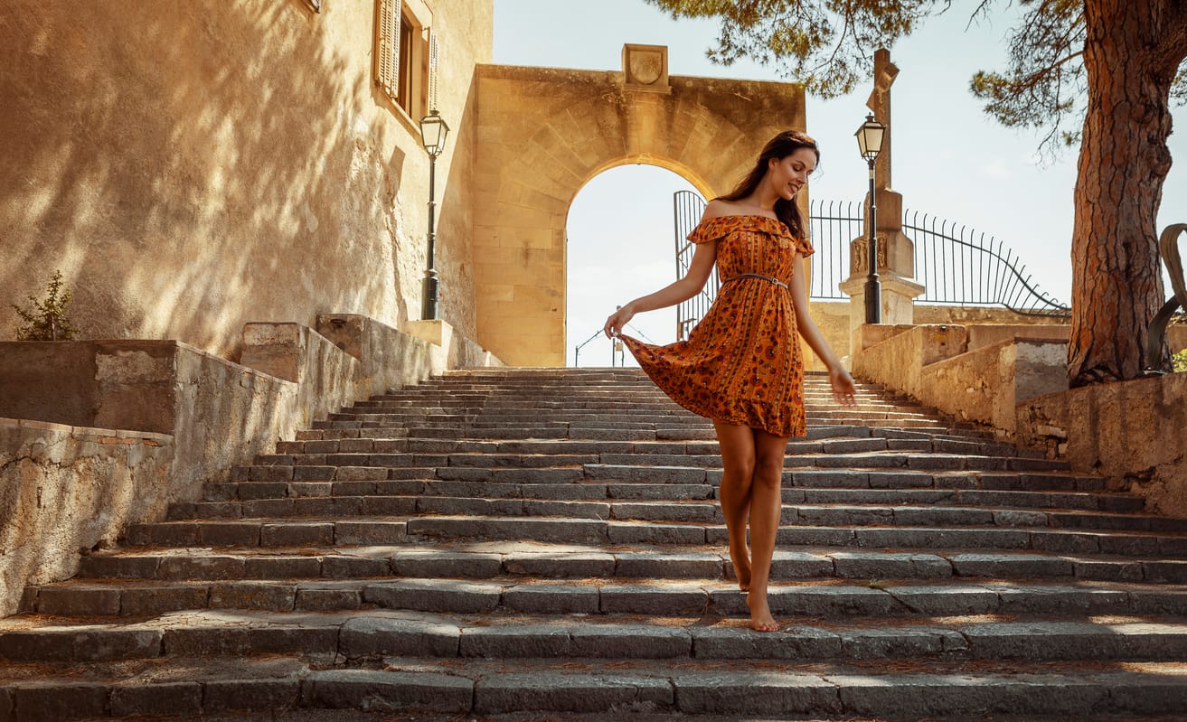 young woman wearing a floral pattern summer dress, walking down a staircase, at a castle in Artà on Mallorca
